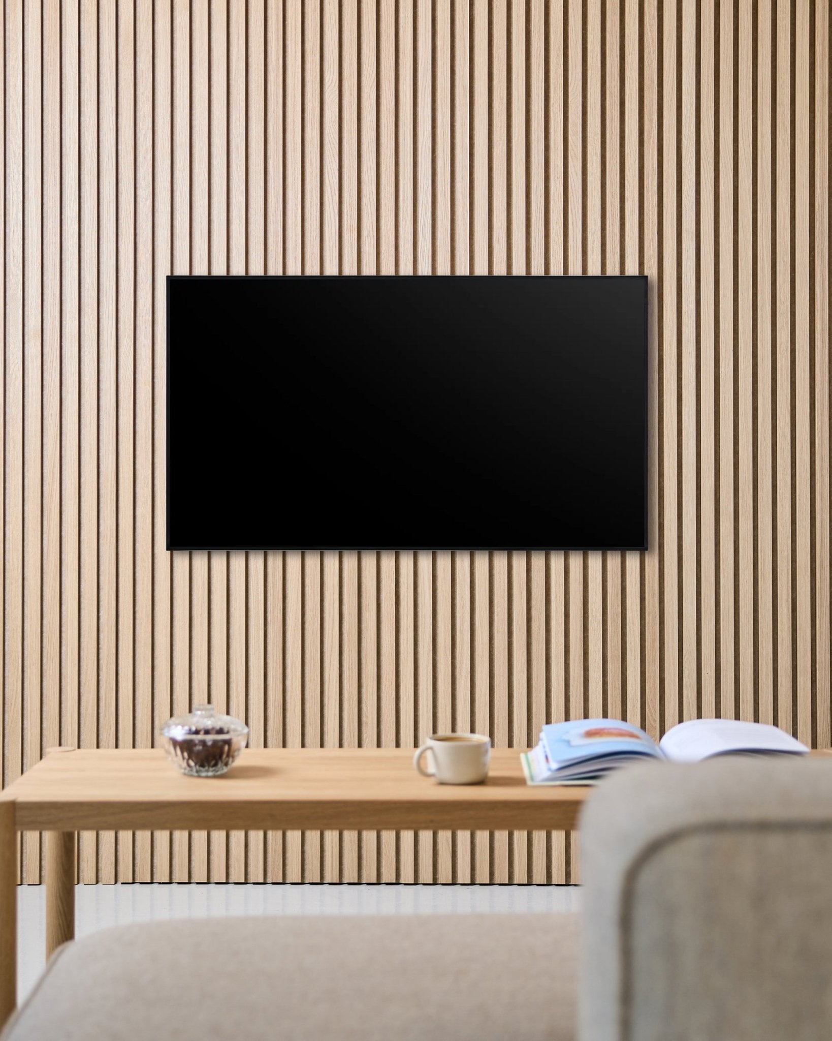 Een Fjell TV Mount (small) van WoodUpp bevestigt een flatscreen aan een Nordische Akupanel akoestische houten lattenwand, boven een tafel met een kopje, een kom en een open tijdschrift naast een beige bank.